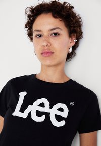 Melna kokvilnas t-kreklu ar lielu balto "Lee" logo. Īsas piedurknes ar apaļu dekoltē un brīvu piegriezumu. Minimāls dizains.