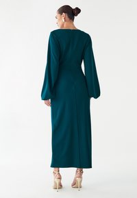 Vestido largo de manga larga en color azul verdoso con cuerpo ajustado, falda fluida y cremallera trasera. Combinado con tacones altos beige con tiras. Tejido suave y ligero.