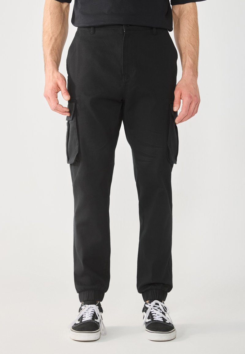 Zwarte cargobroek met taps toelopende enkels, twee zijzakken en een gestructureerde afwerking. Gemaakt met zwart-witte sneakers.