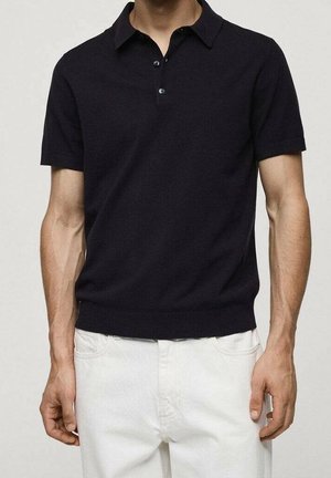 Man draagt een zwart poloshirt met korte mouwen en knopen, gecombineerd met een witte broek, staand tegen een effen achtergrond.