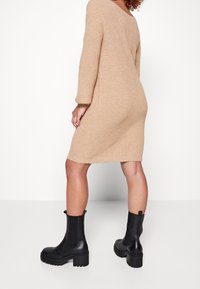 Robe en tricot beige avec des manches longues et une coupe décontractée, associée à des bottines noires à semelle épaisse et panneaux élastiques sur les côtés.