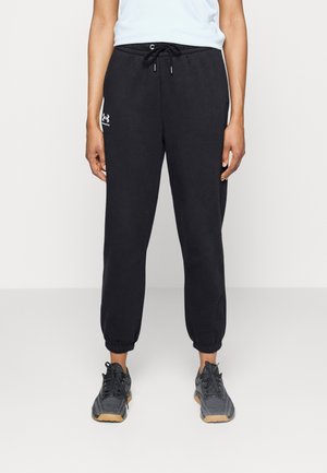 Persoon draagt zwarte Under Armour joggingbroek met elastische boorden en een taillekoord, gecombineerd met zwarte en beige sportschoenen.