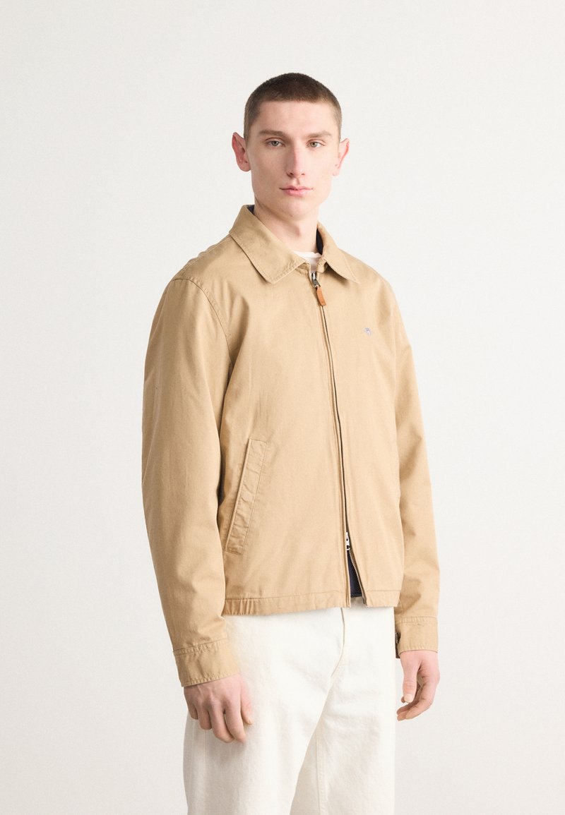 Veste zippée beige avec col large, deux poches latérales et une tirette en cuir, associée à un pantalon blanc, présentée sur fond clair.