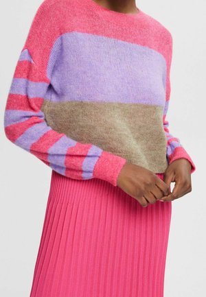 Personne portant un pull color-block avec des sections rose, violette et beige, associé à une jupe plissée rose vif, les mains jointes.
