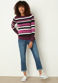 Gestreifter Pullover mit lila, schwarz und weißen Bändern, kombiniert mit hochgekrempelten blauen Jeans und weißen Sneakers. Weiches Strickmaterial.