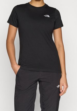 Osoba ubrana w czarną koszulkę z krótkim rękawem i czarne spodnie z logo The North Face na piersi i udzie. Jedna ręka w kieszeni.