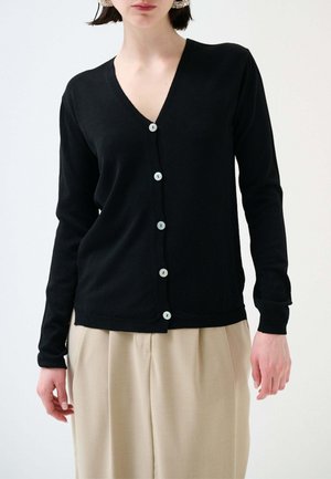 Donna che indossa un cardigan nero a maniche lunghe con bottoni e pantaloni beige plissettati a vita alta, su sfondo uniforme.