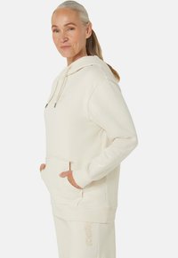 Light beige hoodie made from soft fabric, featuring a front pouch pocket, drawstring hood, and subtle branding on the chest and sleeve.
