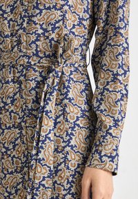 Rochie cu mâneci lungi, de culoare bleumarin, cu un model paisley în nuanțe de portocalie și crem. Are nasturi și o talie marcată cu curea pentru un aspect ajustat.