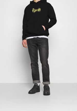 Felpa nera con cappuccio e grafica gialla, abbinata a jeans scuri e sneakers nere. Slim fit, stile casual e polsini arrotolati.