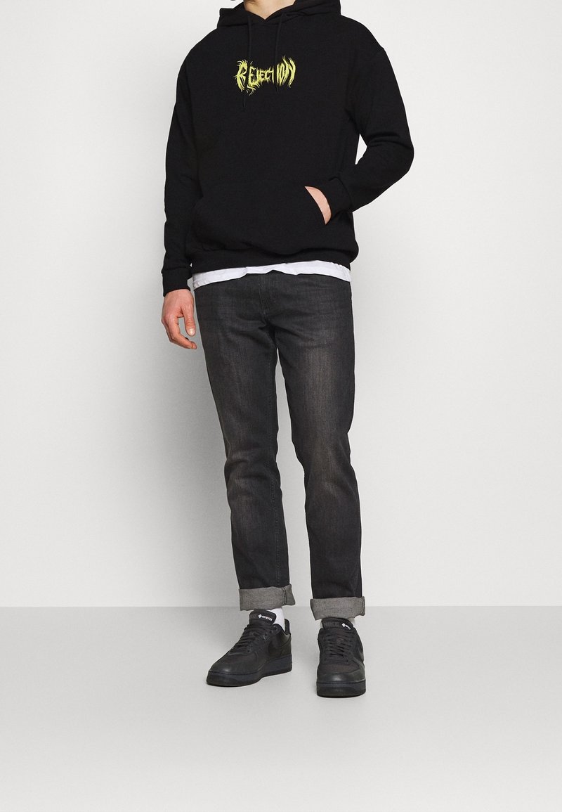 Felpa nera con cappuccio e grafica gialla, abbinata a jeans scuri e sneakers nere. Slim fit, stile casual e polsini arrotolati.