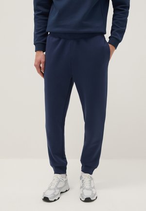 Persoon draagt marineblauwe joggingsbroek en bijpassende trui, gecombineerd met witte sportschoenen, staand tegen een effen achtergrond.