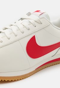 Fehér bőr sportcipő jellegzetes piros Nike plegtetéssel, lapos fűzőkkel és texturált gumi talppal. Minimum dizájn, varrott díszítésekkel.