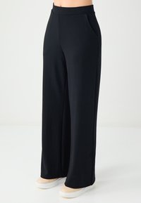 Pantalon large noir en tissu doux et extensible. Doté d'une taille haute et de poches latérales. Design simple et épuré.