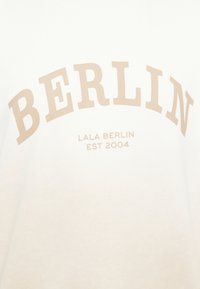 Sweatshirt branco com a palavra "BERLIN" em letras grandes na cor bege clara, e "LALA BERLIN EST 2004" abaixo em fonte menor, feito de material de algodão.