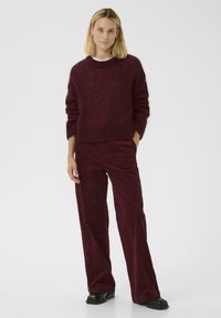 Bordeaux gebreide trui met een ronde hals en geribde manchetten, gecombineerd met bijpassende corduroy broek en zwarte schoenen. Schone achtergrond.