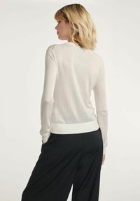 Jasbeżowy sweter z długim rękawem o gładkiej strukturze, z prążkowanymi mankietami i dołem, z przezroczystą tylną częścią. Noszony z czarnymi spodniami.