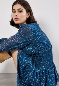 Blau gepunktetes Kleid mit durchsichtigen Ärmeln, gerafftem Oberteil und weit ausgestelltem Rock. Verfügt über weiße Polka-Dots und einen leichten, fließenden Stoff.