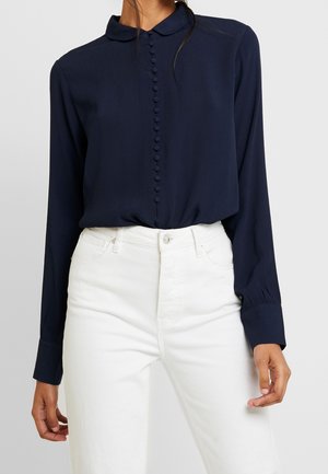 Femme portant un chemisier bleu marine à manches longues avec des boutons recouverts de tissu sur le devant et un pantalon blanc taille haute.