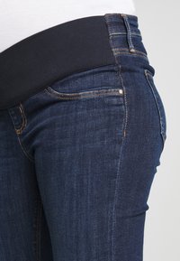 Jeans de mezclilla azul oscuro con una cinturilla elástica negra, que cuentan con un estilo clásico de cinco bolsillos y detalles de costura visibles.