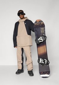 Snieglentininkas vilki smėlio spalvos ir juodą, atsparų orui, striukę ir kelnes, laiko daugiabriaunį snieglenčių su tvirtinimais ir dėvi akinius nuo sniego.