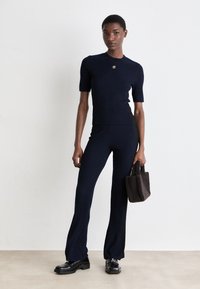 Outfit composto da un maglione a coste blu navy e pantaloni svasati, con collo rotondo, maniche corte e un piccolo dettaglio dorato. Porta una borsa marrone textured.