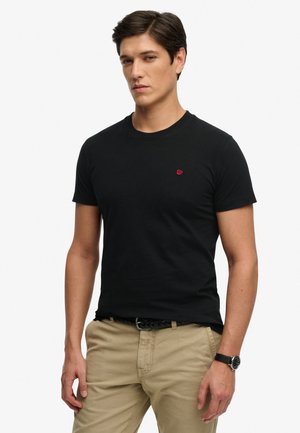 Jeune homme aux cheveux foncés portant un t-shirt noir ajusté avec un logo rouge, un pantalon kaki, une ceinture noire tressée et une montre-bracelet noire sur fond blanc.