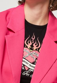 Jasnoróżowa marynarka o strukturalnym fasonie. Pod nią znajduje się czarna koszula z grafiką serca z płomieniami i tekstem.