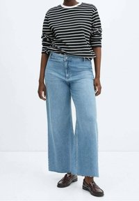 Personne portant un jean large bleu clair, un t-shirt à manches longues rayé noir et blanc, et des mocassins marron foncé, debout sur un fond blanc.