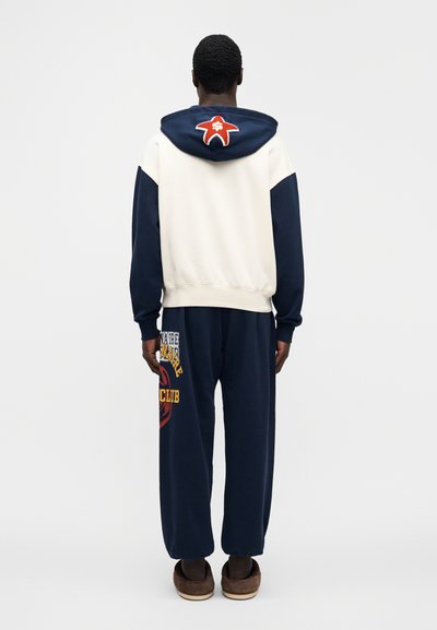 Φούτερ με κουκούλα σε εκρού και μπλε ναυτικό χρώμα με κόκκινο λογότυπο αστεριού στην πλάτη· συνδυασμένο με χαλαρή μπλε ναυτική φόρμα με κίτρινο κείμενο και γραφικό σχέδιο.