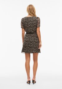 Femme aux longs cheveux blonds portant une robe noire courte à manches courtes, longueur genou, avec des motifs abstraits beiges et des chaussures à talons noirs, debout.