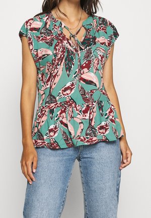 Femme portant un chemisier vert avec un motif de feuilles roses et rouges, un décolleté keyhole et un jean bleu clair.