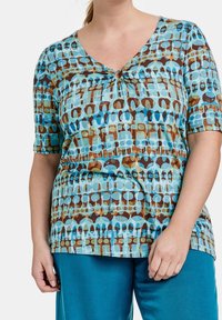 Top a maniche corte in teal con un design a motivi nelle tonalità di blu e marrone, dotato di un dettaglio a chiave sul collo. Tessuto di texture liscia.