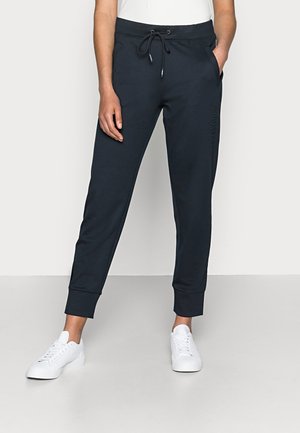 Personne portant un pantalon de jogging noir avec taille à cordon et des baskets blanches, debout avec une main dans la poche.