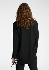 Černý oversized svetr s volným střihem, který má dlouhé rukávy a dekorativní manžety s kontrastním vzorem. Texturovaná látka.