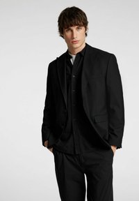 Abito nero con un design slim fit, caratterizzato da un blazer monopetto, rever a scomparsa e una tasca sul petto, indossato sopra una camicia nera a bottoni.