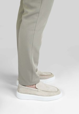 Beige instapsuède schoenen met witte zolen, gedragen bij lichtgrijze, gestructureerde broek tegen een effen achtergrond.