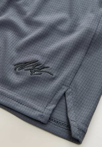 T-shirt de sport gris avec un motif texturé, arborant un logo brodé noir près de l'ourlet et des bords surpiqués, avec des fentes sur les côtés.