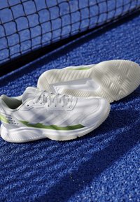 Scarpe da ginnastica bianche con dettagli verdi posizionate su un campo sportivo blu strutturato vicino a una rete nera.