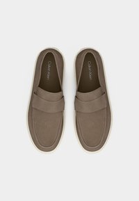 Ζευγάρι από καφέ σουέτ παπούτσια τύπου slip-on με λευκές σόλες, που διαθέτουν φαρδιά λωρίδα στο πάνω μέρος και τη σήμανση "Calvin Klein" στο εσωτερικό.