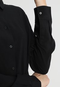 Schwarzes Langarmhemd mit Button-Down-Kragen, ausgestattet mit passenden Knöpfen und strukturiertem Stoff, der ein glattes und maßgeschneidertes Design präsentiert.