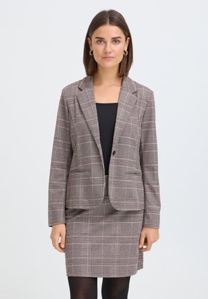 Vrouw met kort bruin haar, gekleed in een bruine geruite blazer en bijpassende rok met zwarte top en zwarte panty's tegen een effen achtergrond.