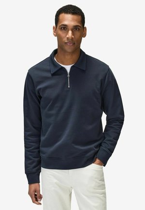 Mørkeblå sweatshirt med lynlås ved kraven, lange ærmer og et struktureret stof. Båret over en hvid skjorte, sammen med lyse bukser.