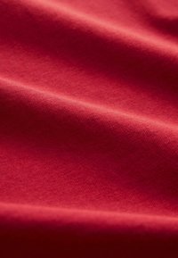 Tissu rouge avec une texture lisse et uniforme ; la lumière se reflète subtilement, créant de doux plis et des ombres à travers la surface, mettant en valeur la profondeur de sa couleur.