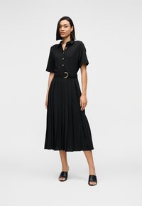 Μαύρο midi φόρεμα πουκάμισο με κοντά μανίκια, χρυσά κουμπιά, ζώνη στη μέση και πλισέ φούστα. Φορεμένο με μαύρα σανδάλια με τακούνι.