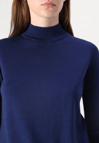 Gros plan d'une personne portant un pull à col roulé ajusté à manches longues de couleur bleu marine avec une texture visible et un col côtelé.