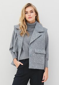 Gray vest with embellished front, large collar, two front pockets, and button closure layered over a gray turtleneck. Black pants visible.