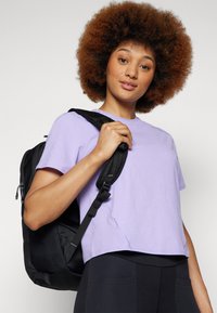 Rucsac negru cu bretele texturate, purtat pe o umăr. Modelul poartă un tricou scurt violet, împreună cu colanți negri cu buzunare.