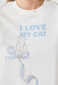 Camiseta de algodón blanca con el texto azul "AMO A MI GATO" y un boceto gris de un gato sentado sobre un lazo de rayas azules.