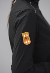 Sort jakke med en gul LED-lyspatch på ærmet. Lyset er ovalformet og har en tænd/sluk-knap samt fire lysende sektioner.